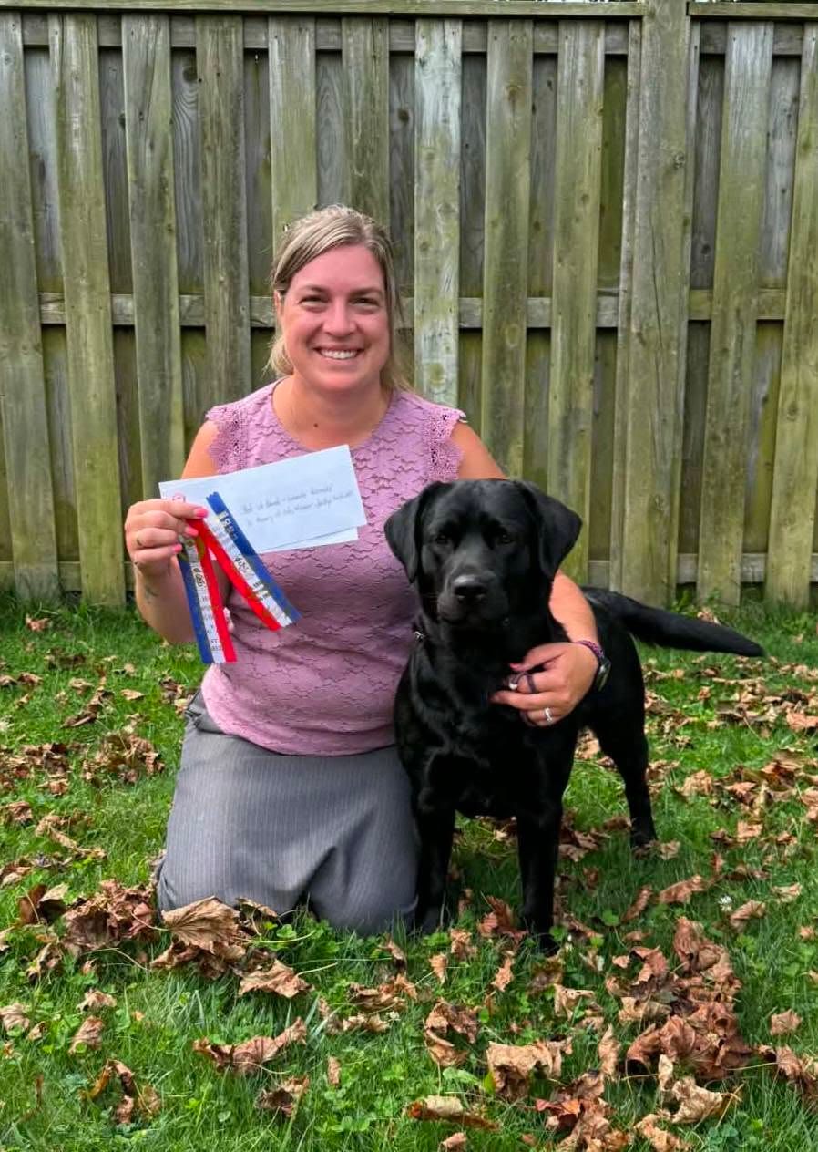 Mandy kneeling with Quill, a black Labrador, holding show ribbons